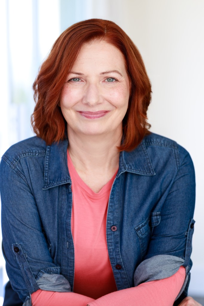 Jennie Olson Six
jennieosix.com
Red haired woman with salmon colored long sleeve shirt, jean shirt layered over the top. 