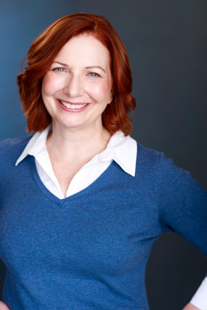 Red haired woman with a white collar shirt and blue sweater. 